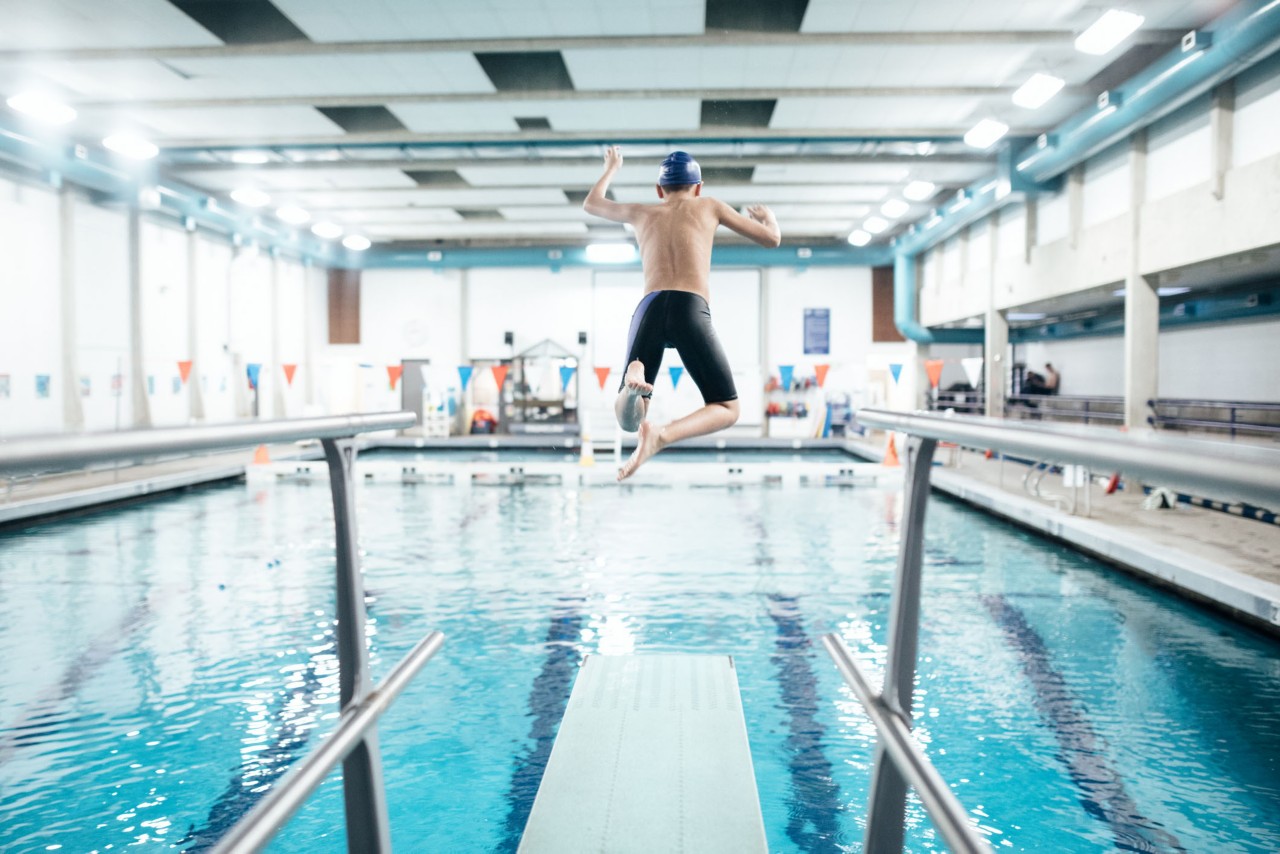 A twelve year old boy enjoys time at the swimming pool jumping off of a diving board into the water.  Healthy active lifestyle that is fun as well.