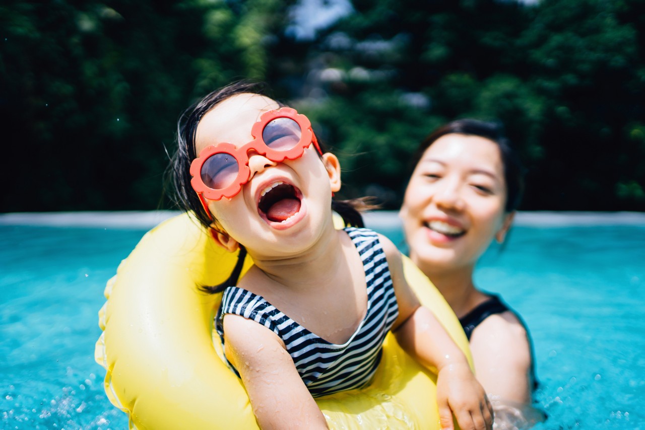Happy toddler girl with sunglasses smiling joyfully, enjoying family bonding time with mother, having fun in the swimming pool in summer
